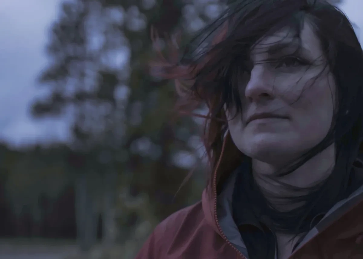 A young woman photographed in a forest at dusk. She is looking pensively beyond the camera, with her hair blowing across her face in the wind 
